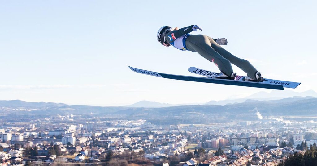 Über den Dächern von Villach: Eine Aufnahme der Skispringerin mitten im Sprung bei hohem Luftstand